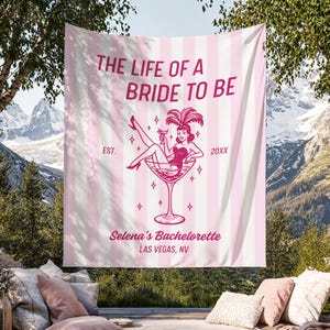 May include: Pink and white striped tapestry with the text "THE LIFE OF A BRIDE TO BE" and "Selena's Bachelorette LAS VEGAS, NV". The design includes a woman in a martini glass. The tapestry is hanging outdoors.