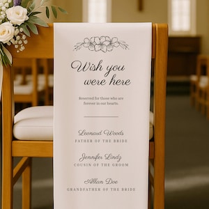 May include: A white fabric memorial sign draped over a wooden chair, featuring a black floral illustration and the text: Wish you were here, Reserved for those who are forever in our hearts, Leonard Woods, Jennifer Lindz, and Allan Doe.