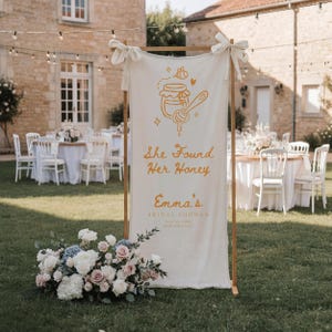 May include: A vertical off-white fabric banner suspended from a wooden frame, featuring a yellow graphic of a honey pot and dipper, with the text "She Found Her Honey", "Emma's", "BRIDAL SHOWER", and "JULY 22, 2024 NEW YORK CITY" printed in yellow.