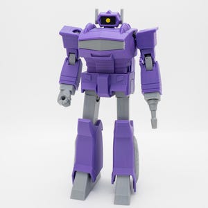May include: A purple robot action figure with gray accents. The toy has a boxy design with articulated arms and legs. The robot's head features a yellow eye and a gray faceplate. The figure stands upright against a white background.