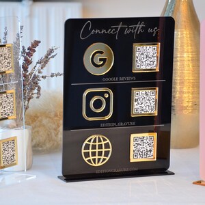 Könnte beinhalten: Acrylschilder mit goldfarbenen Social-Media-Symbolen und QR-Codes. Die Schilder zeigen Google Reviews, Instagram und ein Globus-Symbol. Der Text "Connect with us" steht oben. Die Schilder sind schwarz, transparent und rosa.
