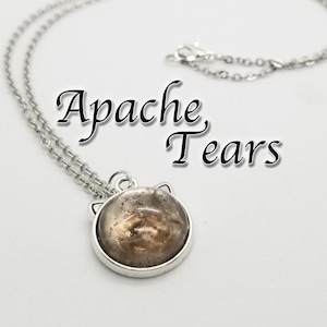May include: A silver chain necklace with a round pendant featuring a brown and clear Apache Tear stone. The pendant has a cat ear design.