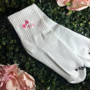 May include: White athletic socks with a pink dumbbell design embroidered on the cuff. The socks are made of a soft material and are suitable for workouts or casual wear. The brand name "KAN" is printed on the sole.