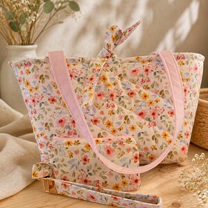 May include: A floral-patterned tote bag, wallet, and wrist strap set. The set features a quilted design with pink, yellow, and blue flowers on a cream background. The tote has pink straps, and the wrist strap has gold-colored hardware.