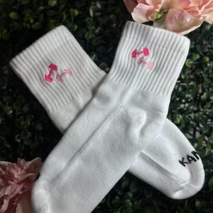 May include: A pair of white athletic socks with a pink dumbbell design embroidered on the cuff. The socks are made of a soft material and are suitable for workouts or casual wear. The word "KAN" is printed on the side of one sock.