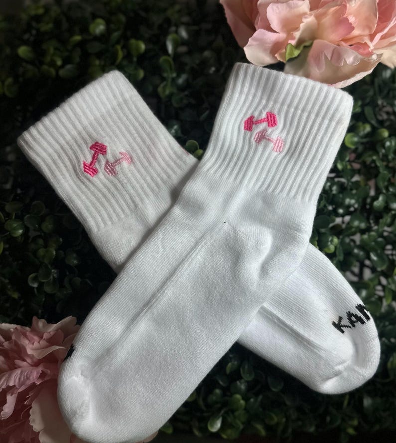 May include: A pair of white athletic socks with a pink dumbbell design embroidered on the cuff. The socks are made of a soft material and are suitable for workouts or casual wear. The word "KAN" is printed on the side of one sock.