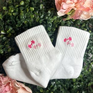 May include: A pair of white ankle socks with a ribbed texture. Each sock features a pink embroidered dumbbell design on the cuff. The socks are displayed against a backdrop of green foliage and pink flowers.