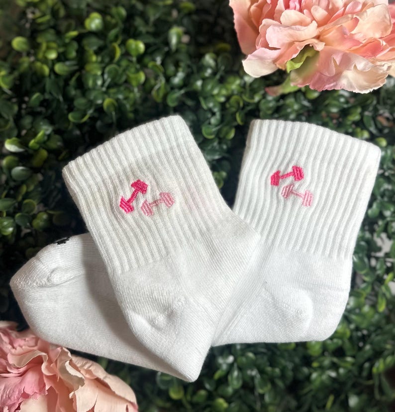 May include: A pair of white ankle socks with a ribbed texture. Each sock features a pink embroidered dumbbell design on the cuff. The socks are displayed against a backdrop of green foliage and pink flowers.