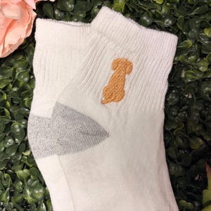May include: A pair of white ankle socks with a light brown embroidered dog design on the cuff. The heel and toe are gray. The socks are made of a soft, ribbed material.