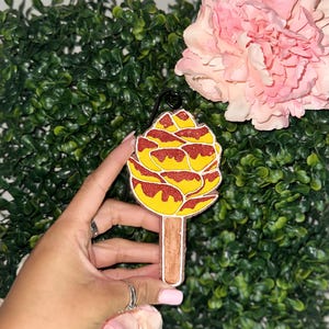 May include: A yellow and red glittery popsicle-shaped ornament with a brown stick. The ornament is held in front of a green leafy background with a pink flower in the upper right corner. The ornament has a black loop at the top.