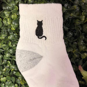 May include: White ankle sock with a black cat silhouette embroidered on the cuff. The sock has a gray heel and toe, and is set against a backdrop of green foliage.