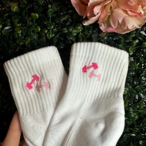 May include: A pair of white athletic socks with a ribbed texture. Each sock features an embroidered pink dumbbell design. The socks are displayed against a backdrop of green foliage and pink flowers.