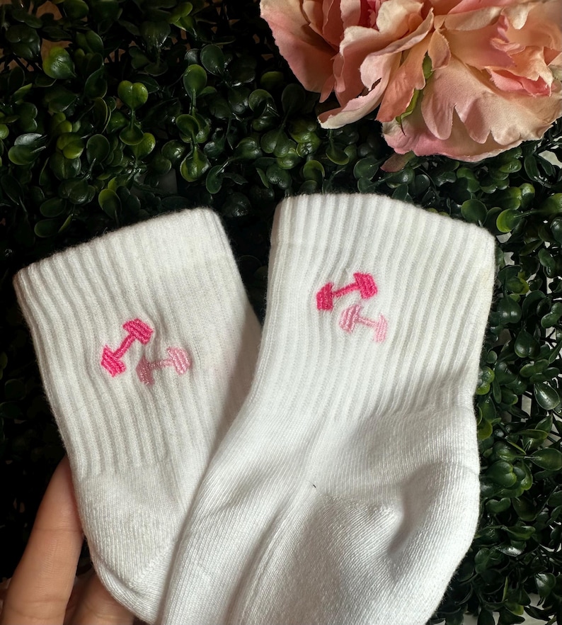May include: A pair of white athletic socks with a ribbed texture. Each sock features an embroidered pink dumbbell design. The socks are displayed against a backdrop of green foliage and pink flowers.