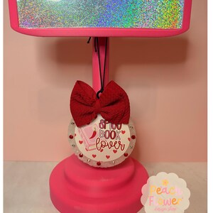 May include: A bright pink T-shaped display stand with a holographic top bar. Hanging from the center is a round white ornament featuring a red bow, small red rhinestones, and the text "SPICY BOOK lover" with a pink book and red chili pepper icon.
