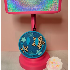 May include: A circular blue glittery smiley face ornament hanging from a bright pink T-shaped display stand. The ornament features two oval eyes, a curved mouth, and several star-shaped accents in solid light blue and leopard print patterns.