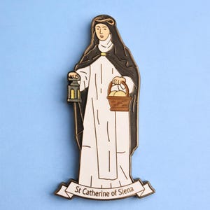 May include: A pin featuring St. Catherine of Siena, depicted in a white robe and black cloak, holding a lantern and a basket. The pin has a gold border and the text "St Catherine of Siena" on a banner.