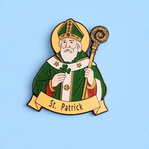 Puede incluir: Un pin de esmalte del Día de San Patricio con una representación de San Patricio con vestiduras verdes, sosteniendo un trébol y un báculo. El pin tiene un halo dorado y una pancarta que dice "St. Patrick".