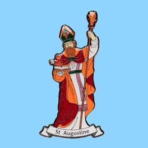Op de afbeelding: Een kleurrijke emaille pin met St. Augustinus, die een staf en boeken vasthoudt. De figuur draagt een rode, oranje en witte jurk, met een groene hoed. De pin heeft een banner aan de onderkant met de tekst "St Augustine".