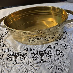 May include: An oval-shaped brass basin featuring a polished gold-tone finish, two side handles, and horizontal ribbed detailing along the exterior walls. The interior base displays a circular embossed stamp mark in the centre of the metal surface.
