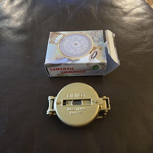 Peut inclure: Une boussole lensatique en métal doré avec un couvercle à charnière portant le texte en relief ENGINEER et DIRECTIONAL COMPASS. La boussole est placée devant son emballage en carton rectangulaire d'origine, qui affiche une image imprimée de la boussole.