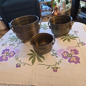 Pode incluir: Um conjunto de três vasos de latão de tamanhos graduados com corpos arredondados, pequenas alças laterais e pés decorativos. Cada vaso apresenta um acabamento metálico liso com uma pátina envelhecida, dispostos sobre uma superfície de tecido branco.
