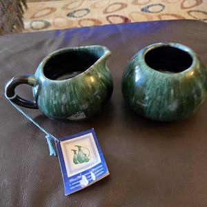 May include: A ceramic creamer and sugar bowl set featuring a distinctive green and black drip glaze finish. The creamer has a curved handle and a pouring spout. A rectangular paper tag is attached, displaying the Blue Mountain Pottery logo.