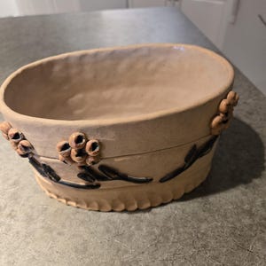May include: An oval-shaped beige ceramic planter with a textured surface. The exterior features raised, sculpted floral accents and dark brown painted vine details. The base of the planter has a scalloped edge design around the bottom perimeter.