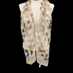 May include: A sheer scarf with a repeating pattern of overlapping circles in shades of brown, beige, and white. The scarf is draped over a mannequin, showcasing its length and design. The edges feature a striped border.