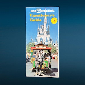 May include: A vintage Walt Disney World Vacationer's Guide magazine cover from July 1980. The cover features a colorful illustration of Cinderella's Castle and a horse-drawn trolley with passengers. The magazine is labeled "Vacationer's Guide" with the number "7" circled in red.