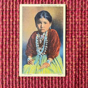 May include: Vintage postcard featuring a portrait of a young girl. She wears a brown long-sleeved top, a turquoise and white beaded necklace, and a striped green and yellow skirt. The text "The Silversmith's Daughter" is at the top.