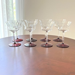 May include: Set of eight clear glass coupe glasses with etched floral designs on the bowl. The glasses have long stems and deep red bases. The glasses are arranged on a round, brown table.