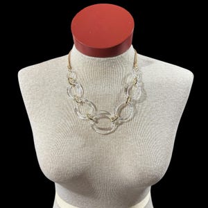 May include: A clear, chunky necklace with gold-tone metal accents. The necklace features large, clear, circular links connected by smaller gold-tone connectors. The necklace is displayed on a mannequin with a red neck and a neutral-colored torso.