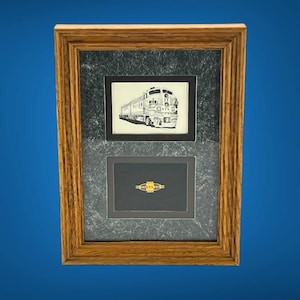 May include: Framed artwork featuring a black and white train drawing above a gold-toned pin with the word "SANTA" on it. The frame is made of wood with a dark gray and black textured matting.