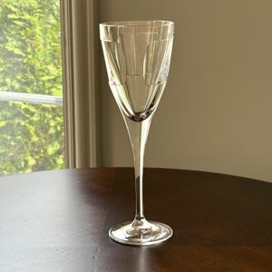 May include: A clear, stemmed glass with a decorative geometric design etched into the upper portion. The glass has a long, slender stem and a wide, circular base. The glass is sitting on a dark brown wooden table.