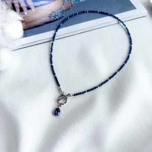May include: A necklace featuring a strand of small, deep blue beads accented with silver-toned spacers. The piece includes a silver-toned toggle clasp and a single teardrop-shaped pendant with a dark blue stone set in a textured silver frame.