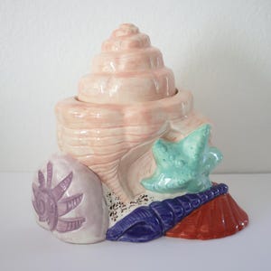 May include: A ceramic seashell-shaped container with a spiral top, painted in shades of pink, purple, blue, and red. A turquoise starfish and other shell details adorn the base. This decorative piece is ideal for coastal decor.