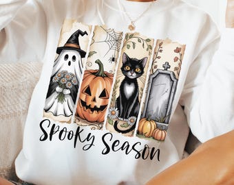 May include: White sweatshirt with a Halloween-themed graphic. The design includes a ghost, jack-o'-lantern, black cat, and tombstone. The words "Spooky Season" are printed below the images. The sweatshirt is a comfortable, casual style.