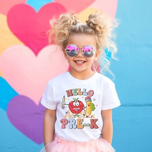 Puede incluir: Una camiseta blanca con un gráfico colorido de una manzana y un lápiz con el texto "HELLO PRE-K". Una niña lleva la camiseta y sonríe.