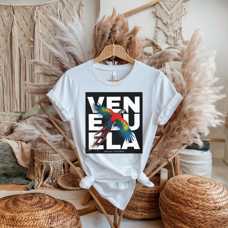 Puede incluir: Camiseta blanca con un gr&aacute;fico cuadrado negro que presenta un loro colorido volando con la palabra "VENEZUELA" en letras blancas y el texto "ABAJO CADENAS" en letras blancas m&aacute;s peque&ntilde;as.
