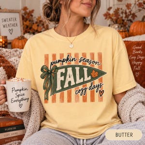Puede incluir: Una camiseta de manga corta de color amarillo pálido con un gráfico de un banderín verde con el texto 'pumpkin season', 'FALL' y 'cozy days' junto a un lazo verde y una hoja de arce. La camiseta se acompaña de una taza blanca con 'Pumpkin Spice Everything'.