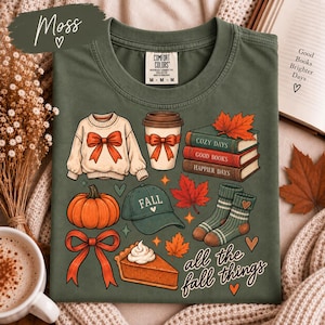 Puede incluir: Una camiseta verde oliva con un estampado gráfico de elementos otoñales que incluye un suéter con lazo, una taza de café, una pila de libros, una calabaza, una gorra con la palabra FALL, calcetines, un trozo de tarta y el texto all the fall things.