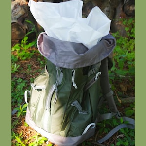 Puede incluir: Una mochila verde y gris con una cubierta de lluvia blanca. La mochila está sentada en un lecho de agujas de pino y plantas verdes.