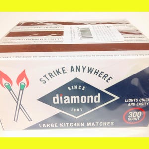 Może przedstawiać: Pudełko z dużymi zapałkami kuchennymi Diamond Strike Anywhere. Pudełko jest białe z brązową górą i zawiera logo diamentu z napisem "Strike Anywhere" i "Lights Quickly and Easily". Pudełko zawiera 300 zapałek.