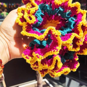 Puede incluir: Una vibrante flor de ganchillo hecha a mano en tonos amarillo, fucsia y turquesa. Los pétalos de la flor están bordeados en amarillo, con una textura arrugada. La artesanía se sostiene frente a un fondo borroso al aire libre.