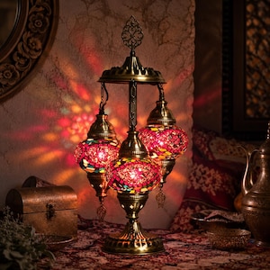 May include: A three-light table lamp featuring a brass-toned metal frame and three spherical glass shades. Each shade is crafted from a mosaic of red, pink, and multi-colored glass pieces that emit a warm, vibrant glow with colorful light patterns.