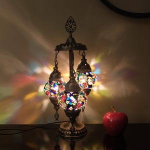 May include: A three-light table lamp with a dark bronze finish and colourful mosaic glass globes. The lamp has a round base and a central stem with three arms that extend out to hold the globes. The globes are made of small pieces of glass that are arranged in a mosaic pattern. The lamp is lit and casting a warm glow.