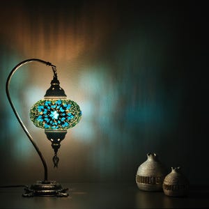 May include: A turquoise and green mosaic glass table lamp with a curved arm and a round base. The lamp is lit and casting a warm glow. Two decorative silver pomegranates are on the table in front of the lamp.