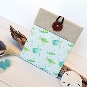 May include: A small, rectangular pouch with a light blue fabric featuring a pattern of green sea turtles. The pouch has a brown button closure and a black elastic band. The pouch is sitting on a piece of driftwood.