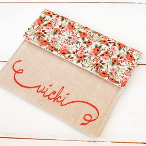 May include: A beige linen clutch with a floral print flap. The clutch has the name "wicki" embroidered in red.