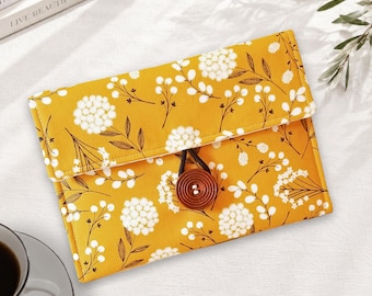 May include: A rectangular fabric pouch in mustard yellow featuring a white and dark brown floral pattern. The pouch has a fold-over flap secured by a dark brown circular wooden button and a thin elastic loop closure centered on the front.